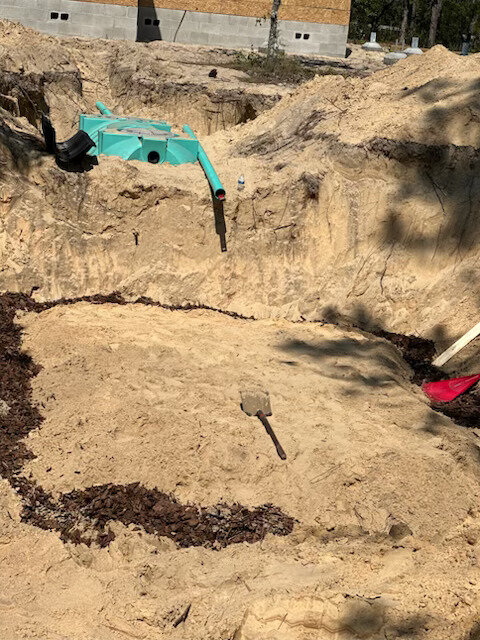 Excavated drain field bed in sandy soil with a septic tank and green PVC pipe at the edge of the dig