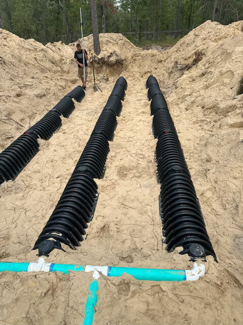 Three rows of black plastic drain field chambers laid in an excavated sand bed, joined by a PVC distribution line